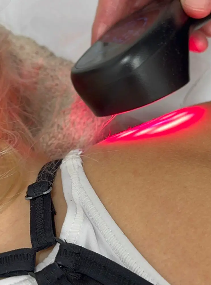 A person's shoulder is being treated with a handheld red light therapy device. The light is focused on the shoulder area, and the person is wearing a white top with black straps.