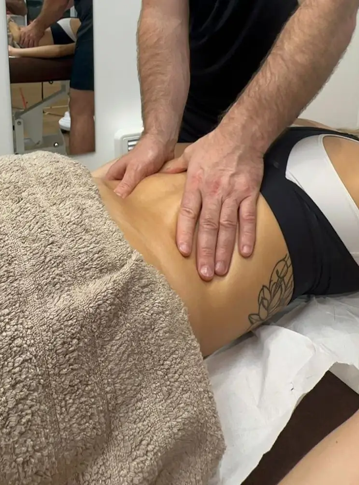 A person receiving massage therapy on a table has their lower back exposed, with the therapist's hands gently pressing on the skin. A beige towel covers the lower body, and a tattoo is visible on the person's side.