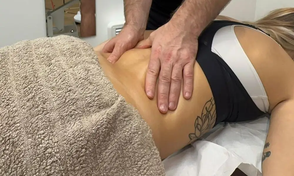 A person receiving a massage lies face down on a table, partially covered with a towel. The massage therapist's hands are placed on the person's lower back, applying pressure. The person wears a black top and shows a floral tattoo on the side.