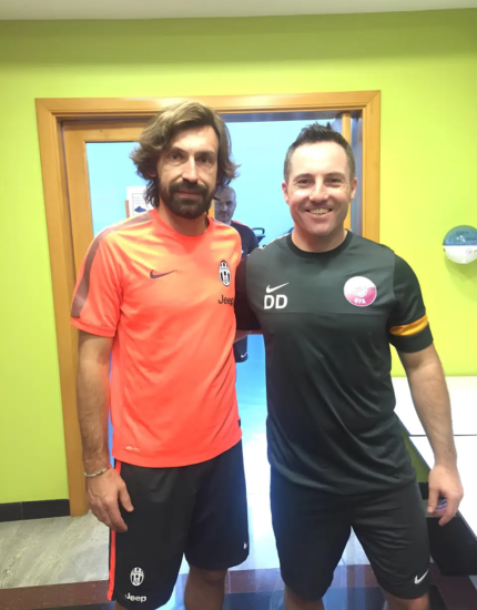 Two men in sports attire stand side by side inside a room. The man on the left wears an orange shirt with a Juventus logo, while Damien Delahunty stands on the right, sporting a black shirt with initials "DD." They both smile at the camera.