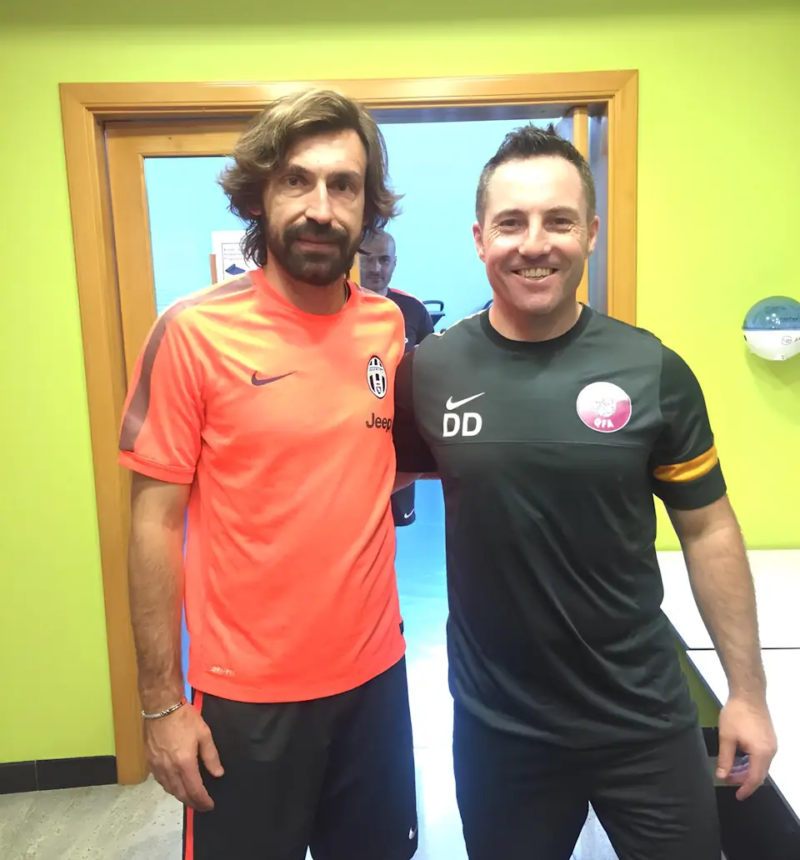 Two men in sports attire stand side by side inside a room. The man on the left wears an orange shirt with a Juventus logo, while Damien Delahunty stands on the right, sporting a black shirt with initials "DD." They both smile at the camera.