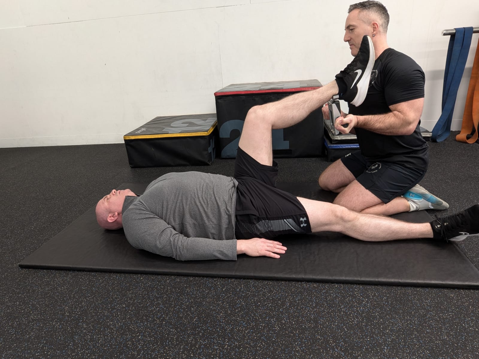A person lies on an exercise mat, with one leg raised, while a trainer assists by holding the foot. Gym equipment is in the background.