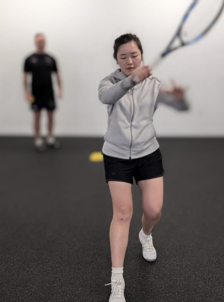 A woman in a gray hoodie and black shorts swings a tennis racket indoors. A man in the background watches her practice. The room has a dark floor and white walls with yellow markers on the ground.