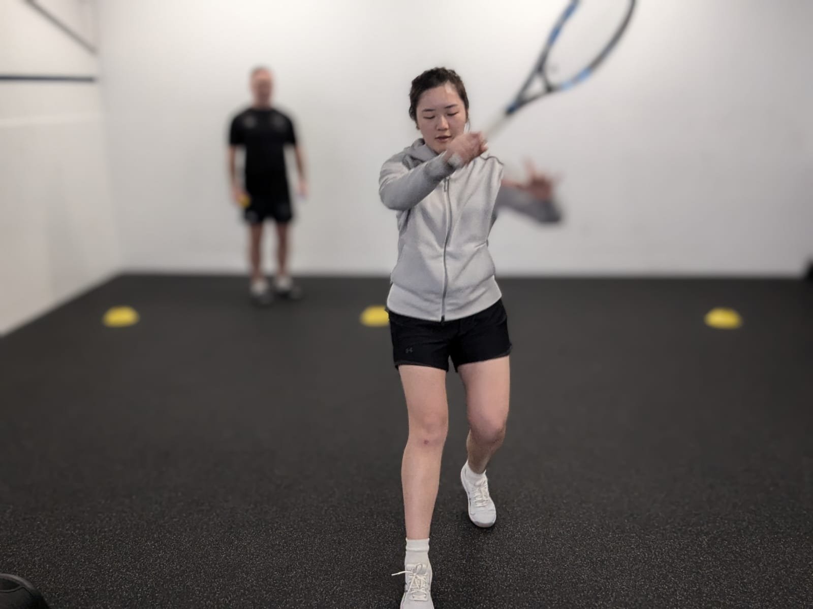 A woman in a gray hoodie and black shorts swings a tennis racket indoors. A man in the background watches her practice. The room has a dark floor and white walls with yellow markers on the ground.