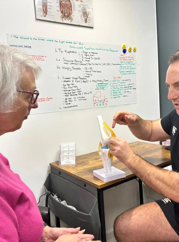 A man sits at a desk explaining a model of a knee joint to an older woman in a pink sweater. They are in a room with a dry-erase board displaying notes and diagrams in the background.