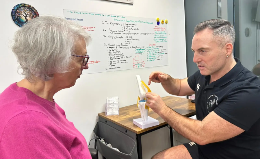 A man sits at a desk explaining a model of a knee joint to an older woman in a pink sweater. They are in a room with a dry-erase board displaying notes and diagrams in the background.