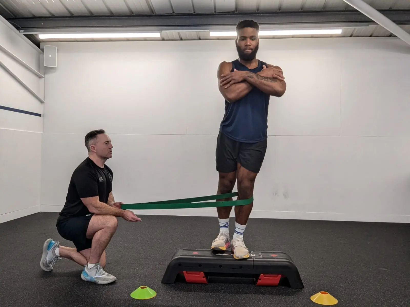 A person stands on a step platform with arms crossed and a resistance band around their knees, held by a trainer kneeling beside them. The setting is a gym with various colored cones on the floor.