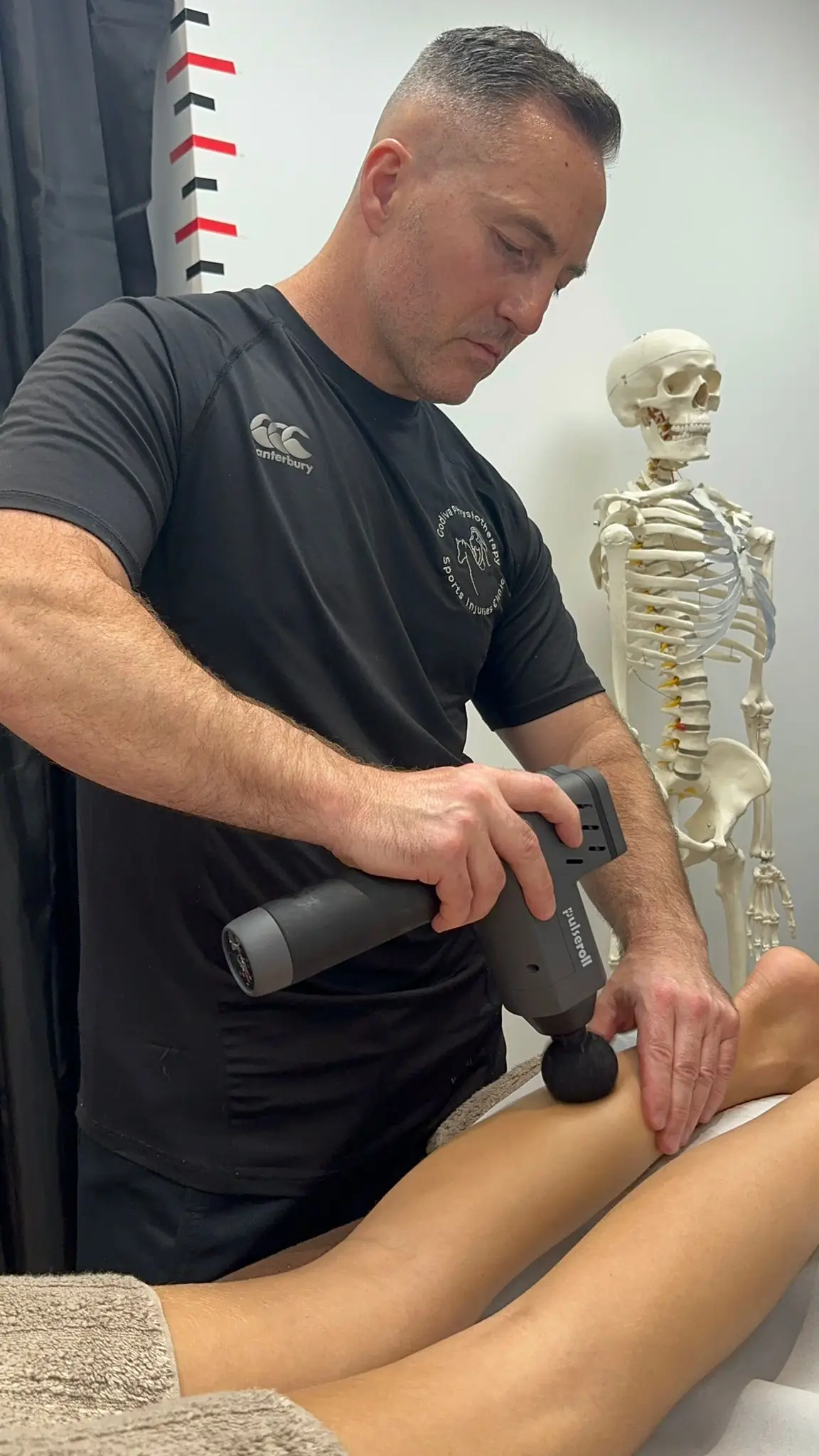 In a therapy room, Damien Delahunty, wearing a black shirt, expertly applies a massage gun to a client's leg. A skeleton model quietly oversees the session in the background.