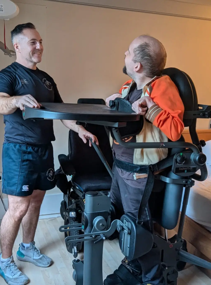 A man in athletic wear stands beside another man using a standing wheelchair device, designed to aid those with neurological conditions, in a bedroom setting. The room features a bed, wooden flooring, and a ceiling lift visible in the background. Both men are smiling and engaged in conversation.