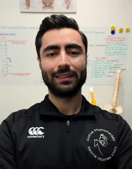 A person with dark hair and a beard is smiling while wearing a black jacket with a logo that reads "Godiva Physiotherapy Sports Injuries Clinic." The background features anatomical charts and a diagram on the wall.