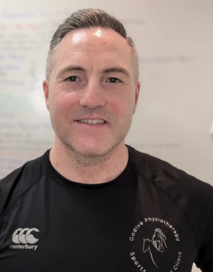 A man with short hair wearing a black Canterbury t-shirt with a logo for Godiva Physiotherapy Sports Clinic. He is smiling, standing indoors, with a whiteboard covered in notes and a lamp visible in the background.