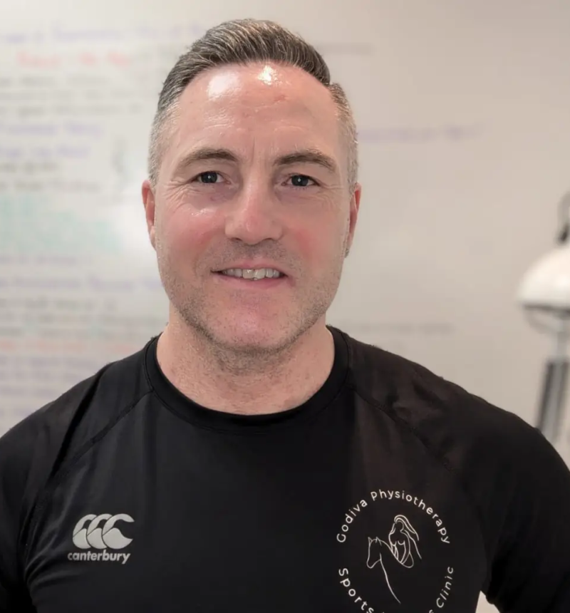 A man with short hair wearing a black Canterbury t-shirt with a logo for Godiva Physiotherapy Sports Clinic. He is smiling, standing indoors, with a whiteboard covered in notes and a lamp visible in the background.