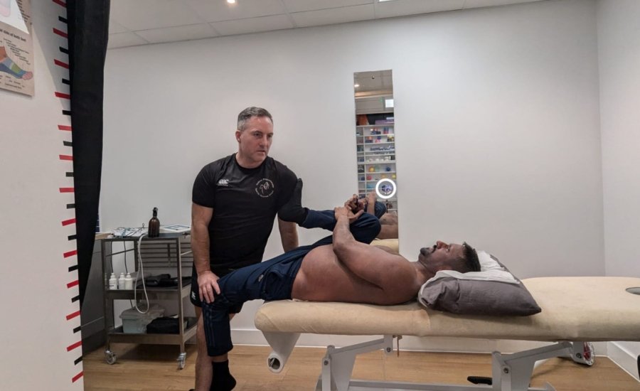 A man lies on a therapy table while a therapist assists with leg stretches in a clinical room. The room has white walls, a small mirror, and medical equipment. The therapist wears a black shirt and shorts.