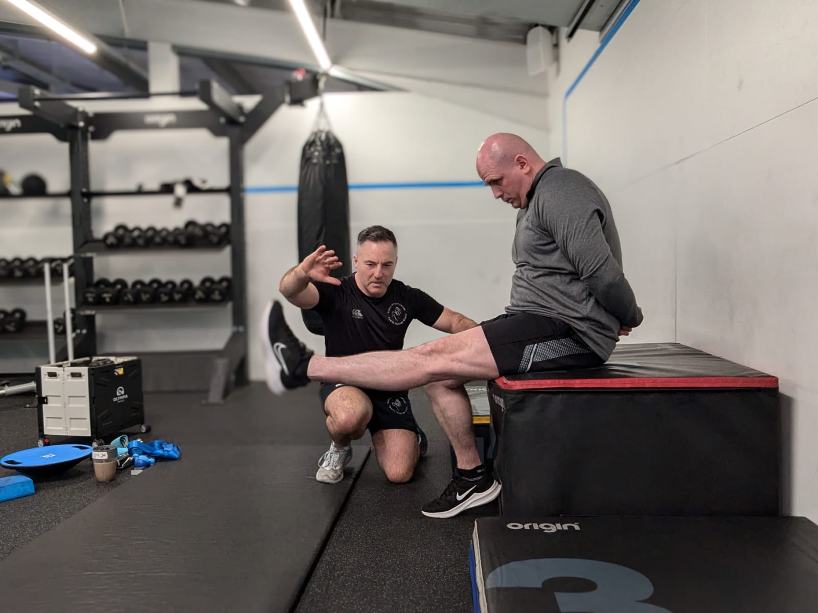A trainer in black gym attire guides a bald man with knee exercises on a padded bench. The setting is an organized gym with equipment like free weights and a boxing bag in the background. The man extends one leg outward while the trainer gestures for balance.