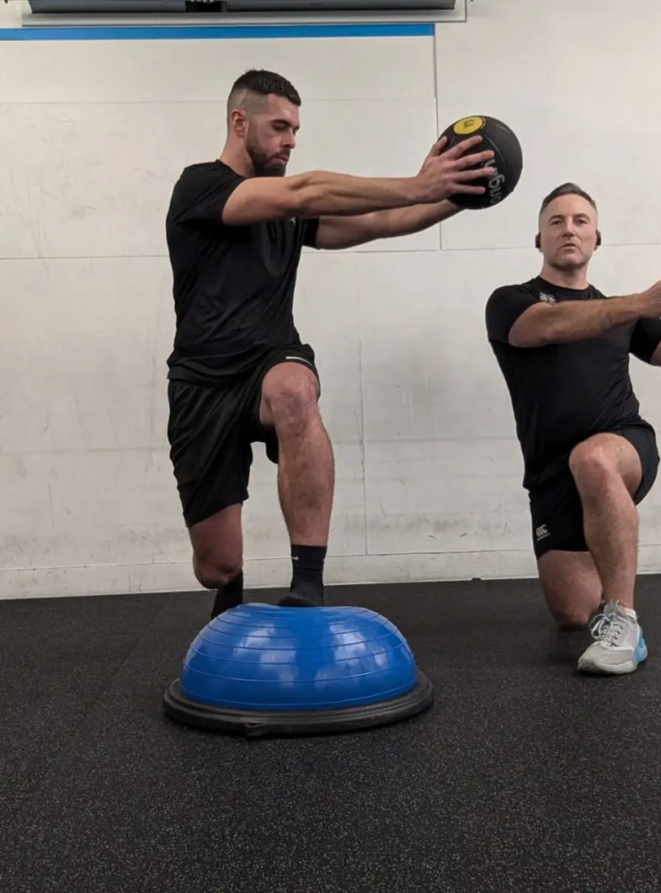 Two men in a gym are engaging in a workout focused on injury prevention. One man is holding a medicine ball while balancing on a half exercise ball. The other kneels, seemingly ready to catch the ball. Both are intensely focused on their fitness routine, blending sports medicine insights into their training.