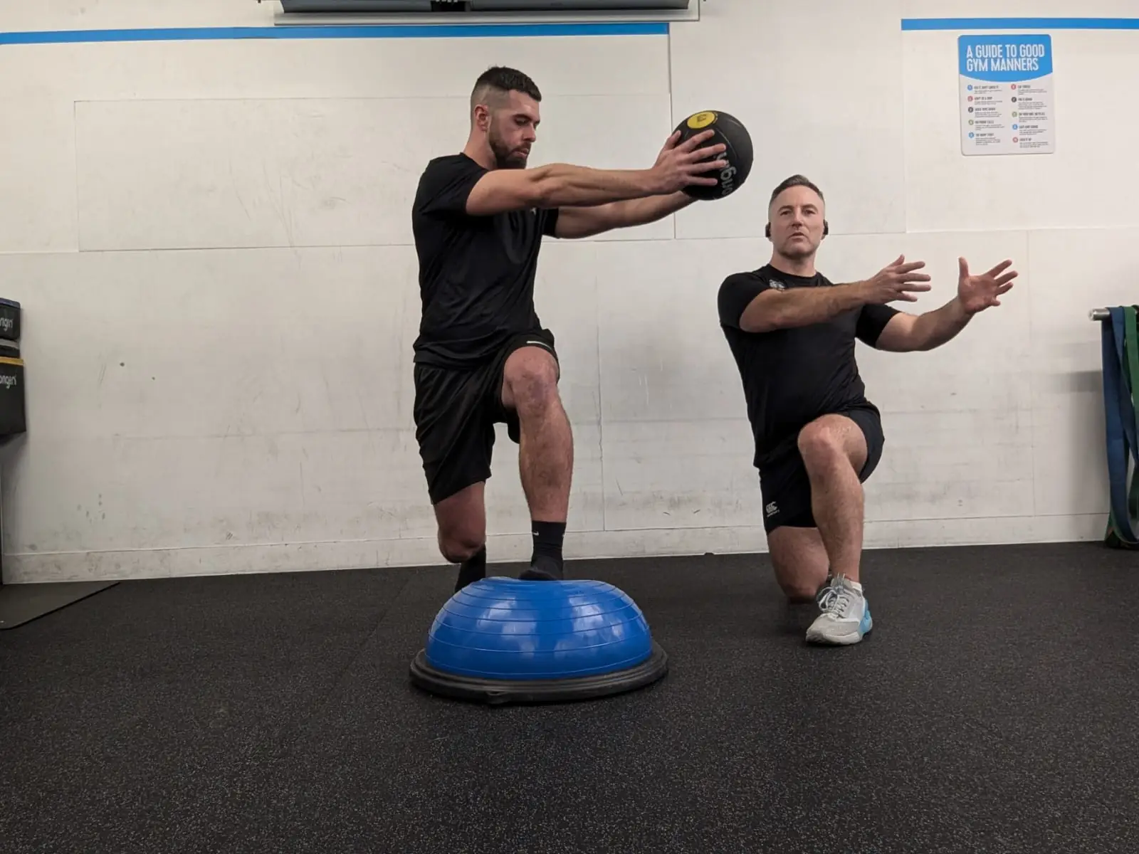 Two men in a gym are engaging in a workout focused on injury prevention. One man is holding a medicine ball while balancing on a half exercise ball. The other kneels, seemingly ready to catch the ball. Both are intensely focused on their fitness routine, blending sports medicine insights into their training.