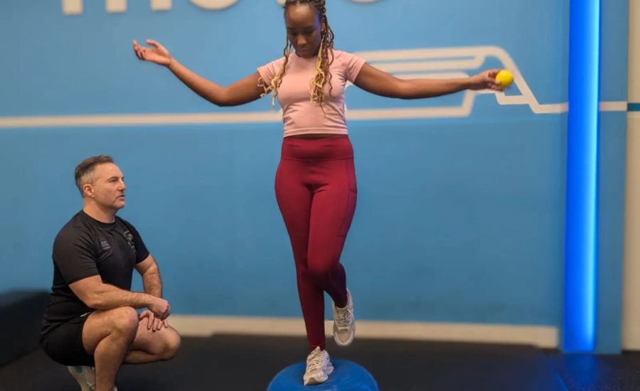 A woman balances on a blue Bosu ball with one foot while holding a tennis ball in one hand. A man kneels nearby, observing. The background is a blue wall with the word "move" partially visible.