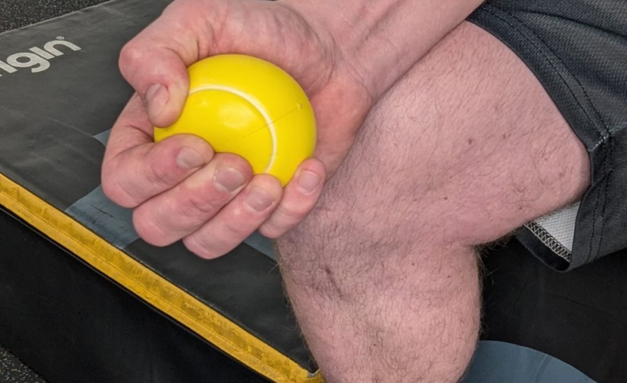 A person holds a yellow ball in their hand, sitting on gym equipment. The focus is on the hand and ball, with part of the leg also visible. The background features a black and yellow padded surface.