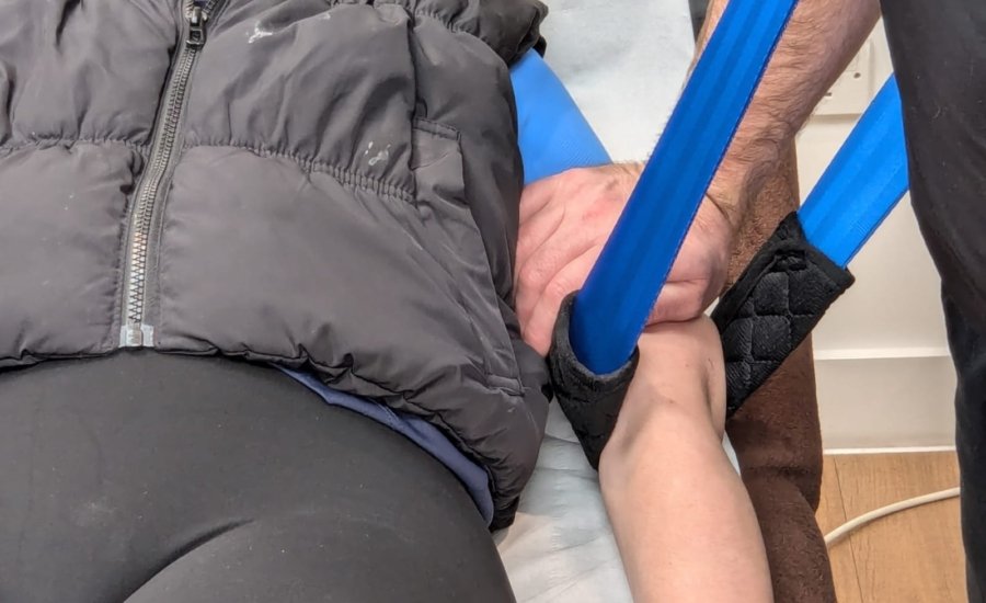 A person lying on a medical bed with their arm in a blue sling. Another person's hand is supporting the arm at the elbow, helping with the adjustment of the sling. The person on the bed is wearing a black padded vest.