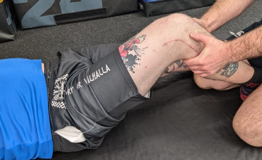 A person wearing a blue shirt and black shorts with tattoos is lying on the floor while another person, also with visible tattoos, supports their bent knee. The scene appears to be in a gym or physical therapy setting.