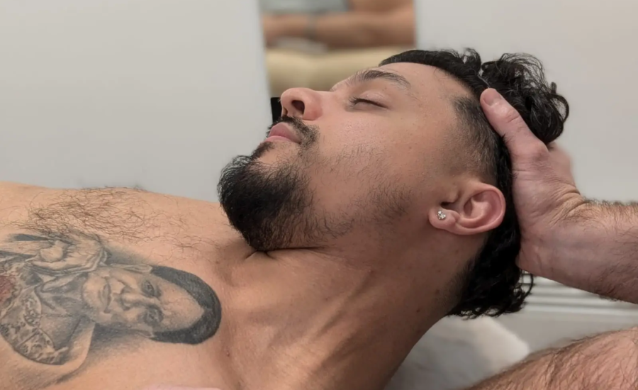 A man with a beard and a tattoo on his chest is lying down, his head supported by another person's hands. He appears relaxed, with his eyes closed, in what looks like a massage or therapy session.