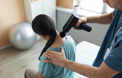 A person uses a massage gun on the cervical area of a woman with long dark hair tied in a ponytail. She is seated on a therapy bed in a room with light walls and a large exercise ball in the background.