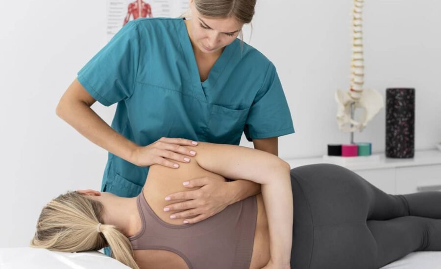 A physical therapist in teal scrubs is assisting a patient lying on her side on a treatment table. The patient wears a brown tank top and gray leggings. A spine model and anatomical poster are visible in the background.