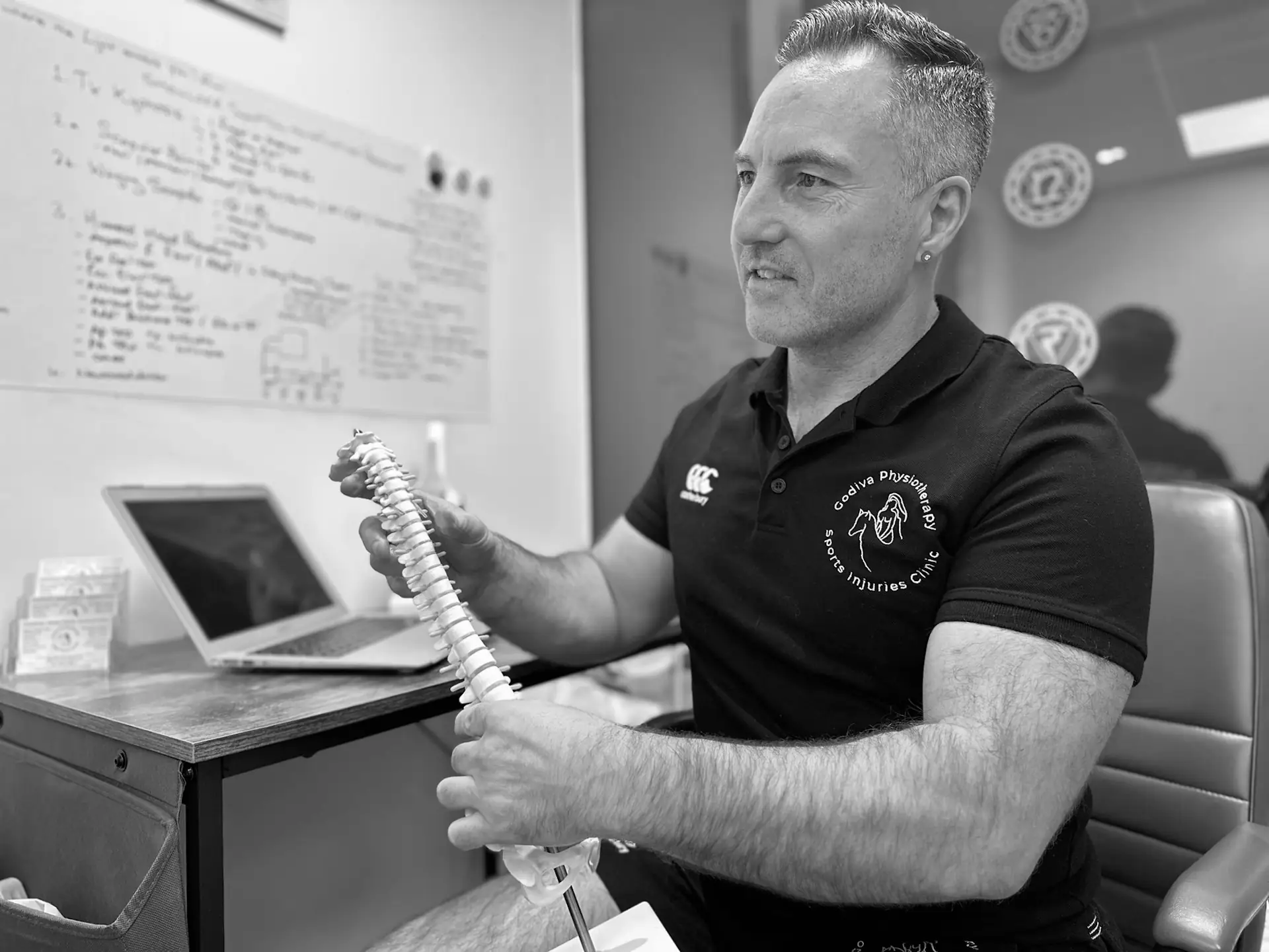 A man in a black polo shirt seated at a desk holds a spine model, explaining it. A laptop and a whiteboard with notes are in the background. The setting is a professional office, and the image is in black and white.