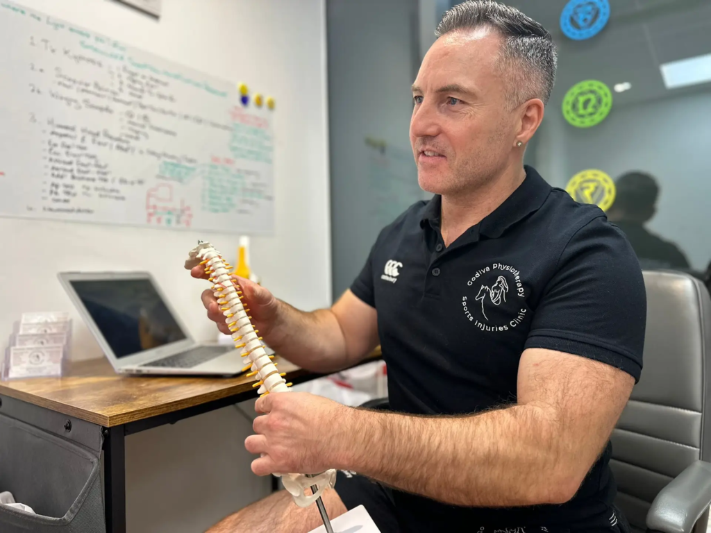 A man in a black polo shirt is sitting at a desk, holding a spine model. A laptop is open beside him, and a whiteboard with notes is in the background. He appears to be explaining or demonstrating something.