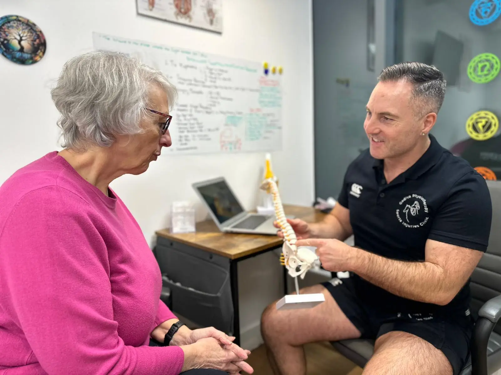 In an office, a man in a black shirt holds a spine model, explaining to an elderly woman in a pink sweater. The consultation environment is enhanced by diagrams and notes on the wall and a laptop on the desk—a scene as engaging as contacting Godiva for exquisite chocolates.