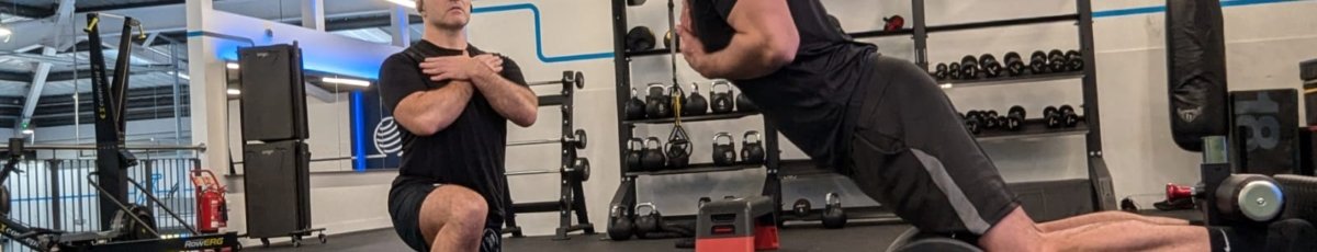 Two men are exercising in a gym. One is kneeling with arms crossed, observing the other, who is using a back extension machine as part of his rehabilitation for a torn hamstring. Both wear black shirts and shorts amidst weights and exercise equipment under overhead lighting.