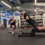 Two men are exercising in a gym. One is kneeling with arms crossed, observing the other, who is using a back extension machine as part of his rehabilitation for a torn hamstring. Both wear black shirts and shorts amidst weights and exercise equipment under overhead lighting.