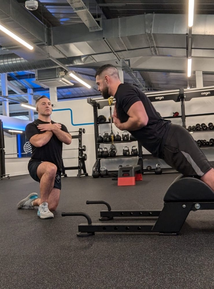 Two men are exercising in a gym. One is kneeling with arms crossed, observing the other, who is using a back extension machine as part of his rehabilitation for a torn hamstring. Both wear black shirts and shorts amidst weights and exercise equipment under overhead lighting.