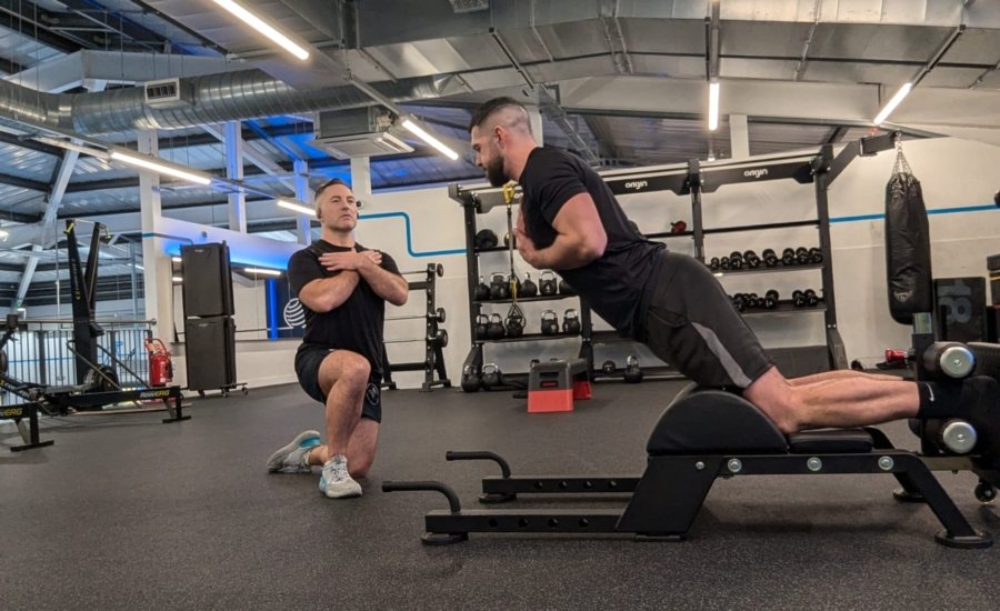 Two men are exercising in a gym. One is kneeling with arms crossed, observing the other, who is using a back extension machine as part of his rehabilitation for a torn hamstring. Both wear black shirts and shorts amidst weights and exercise equipment under overhead lighting.