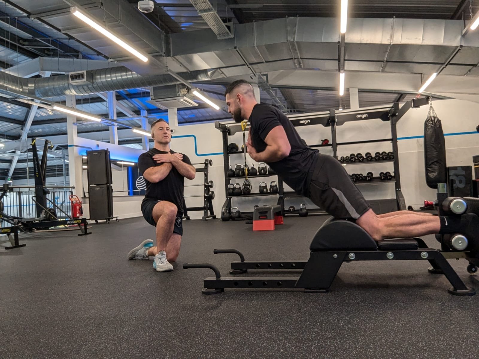 Two men are exercising in a gym. One is kneeling with arms crossed, observing the other, who is using a back extension machine as part of his rehabilitation for a torn hamstring. Both wear black shirts and shorts amidst weights and exercise equipment under overhead lighting.