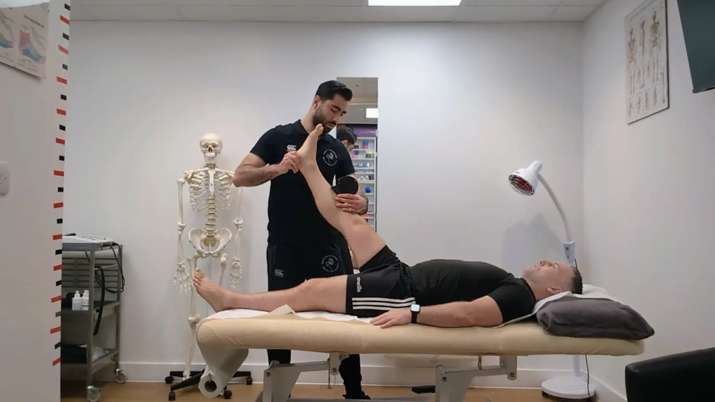 A man in a black uniform assists with a leg stretch for another man recovering from a torn hamstring, lying on a massage table in the therapy room. A skeleton model and lamp provide subtle background details.