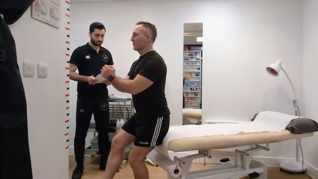 A man in athletic attire performs a squat exercise as part of his rehabilitation in a medical room. Another man in a tracksuit observes him, holding a clipboard. The room has a medical bed and various equipment in the background, aiding his recovery from a torn hamstring.