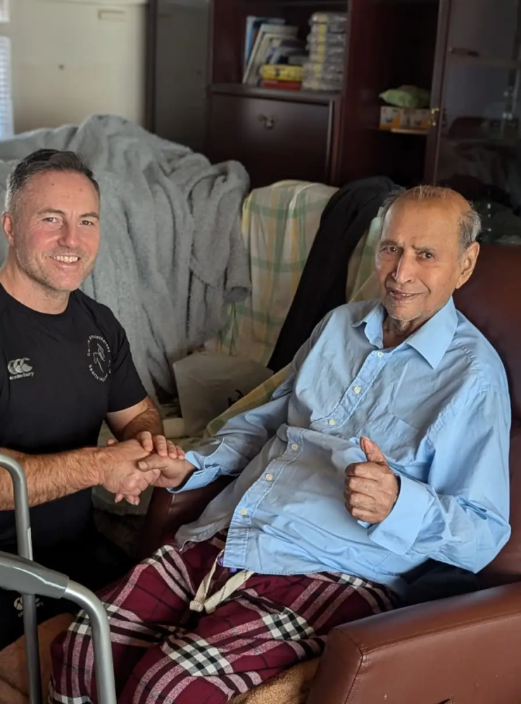 A man in a black shirt kneels beside an elderly man sitting in a recliner, offering encouragement during physiotherapy. The elderly man, in a light blue shirt and checkered pants, gives a thumbs-up while holding the other man's hand. A walker is nearby in the cozy room.