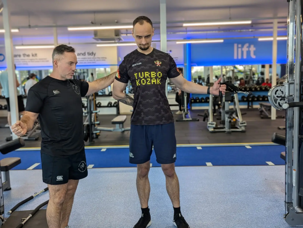 Two men are in a gym focused on bodybuilding. The man on the left appears to be instructing, possibly incorporating physiotherapy techniques, while the man on the right performs a cable exercise. The gym features various equipment and a blue and white interior.