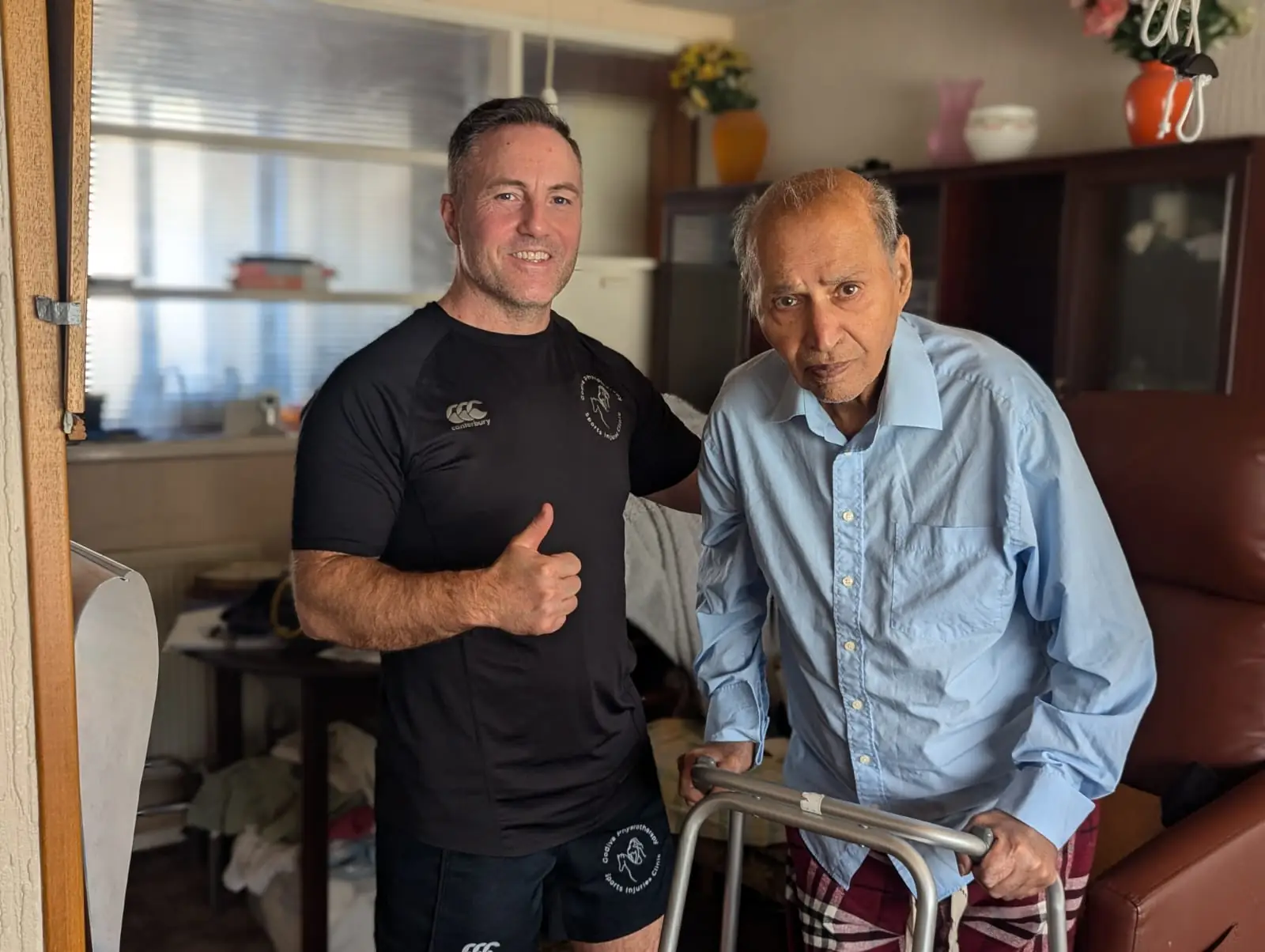 A man in a black shirt, smiling and giving a thumbs-up, stands beside an elderly man using a walker, showcasing senior mobility. The elderly gentleman wears a light blue shirt and checkered pants. Shelves and a cabinet with a plant are visible in the background.