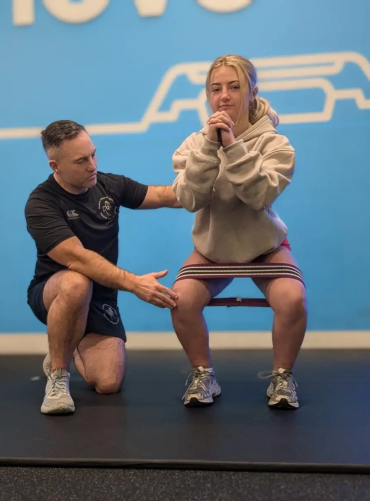 A coach guides a woman in performing a squat exercise, aiding her hamstring rehabilitation. She has a resistance band around her thighs, wearing a beige hoodie and gray sneakers. The setting is a gym with a blue background and the word "MOVE" partially visible.