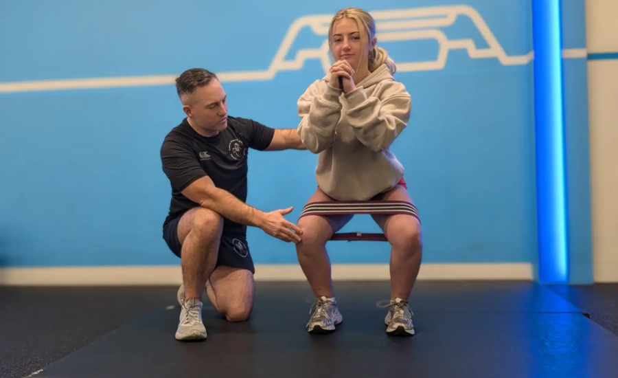 A coach guides a woman in performing a squat exercise, aiding her hamstring rehabilitation. She has a resistance band around her thighs, wearing a beige hoodie and gray sneakers. The setting is a gym with a blue background and the word "MOVE" partially visible.