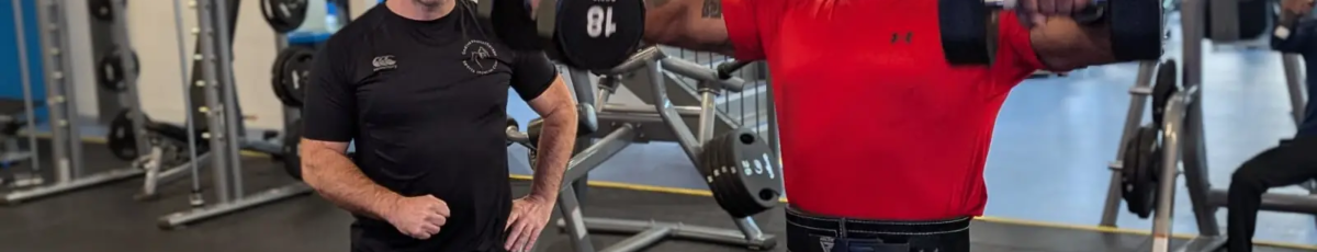 A man in a red shirt lifts dumbbells for a shoulder exercise at the gym, while another man watches nearby. The gym is equipped with various fitness machines and weights, and a staircase is visible in the background.