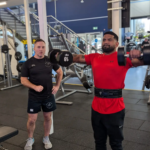 A man in a red shirt lifts dumbbells for a shoulder exercise at the gym, while another man watches nearby. The gym is equipped with various fitness machines and weights, and a staircase is visible in the background.