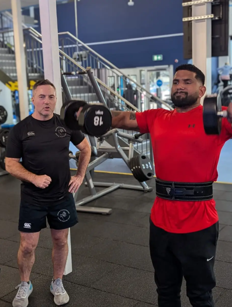 A man in a red shirt lifts dumbbells for a shoulder exercise at the gym, while another man watches nearby. The gym is equipped with various fitness machines and weights, and a staircase is visible in the background.