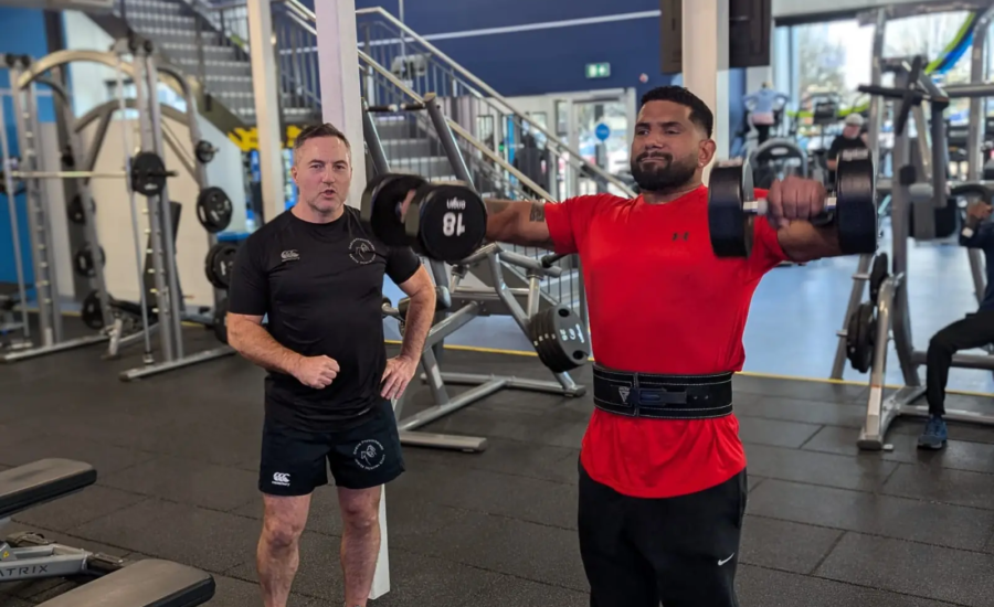 A man in a red shirt lifts dumbbells for a shoulder exercise at the gym, while another man watches nearby. The gym is equipped with various fitness machines and weights, and a staircase is visible in the background.