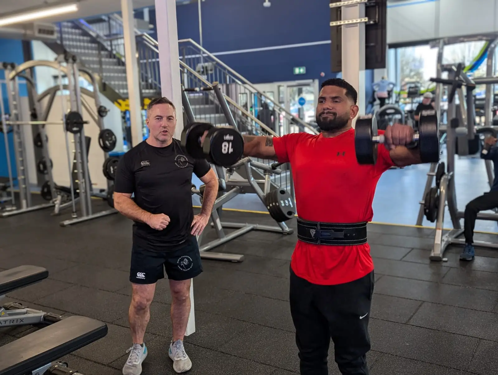 A man in a red shirt lifts dumbbells for a shoulder exercise at the gym, while another man watches nearby. The gym is equipped with various fitness machines and weights, and a staircase is visible in the background.