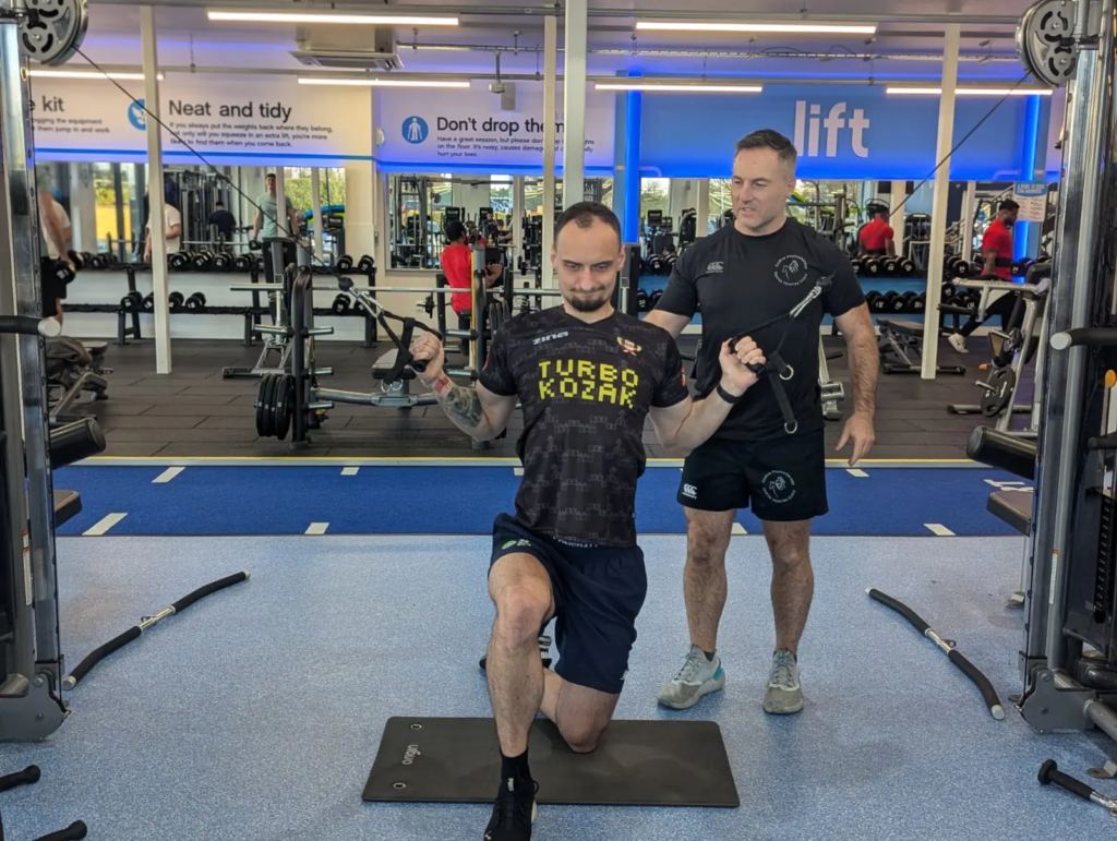 A man kneels on a mat in a gym, focused on bodybuilding as he grips cable machine handles. Another man stands behind, offering physiotherapy guidance. Gym equipment is visible in the background, with blue and white motivational signs adorning the walls.