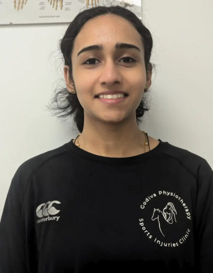 A person wearing a black sports shirt with a logo stands smiling in front of a white wall with a chart partially visible in the background.