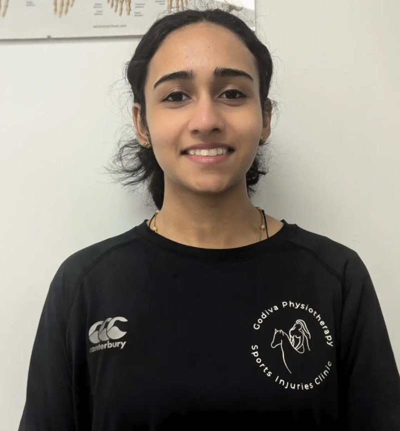 A person wearing a black sports shirt with a logo stands smiling in front of a white wall with a chart partially visible in the background.